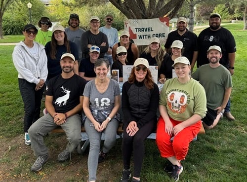A group of 18 men and women pose for a photo in a park-like setting beneath a banner that reads Travel With Care.