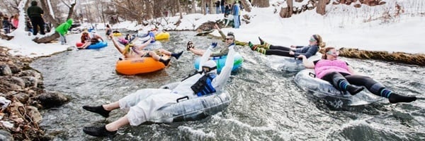 costumed men and women float down in a river on colorful tubes on a snowy day, as part of a winter festival.